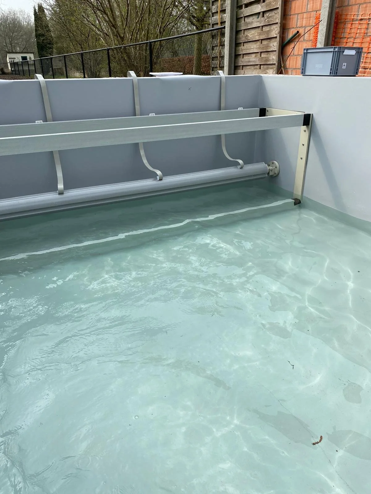 Een leeg zwembad met water en een metalen installatie aan de muur, omgeven door een afrastering en een houten hek.