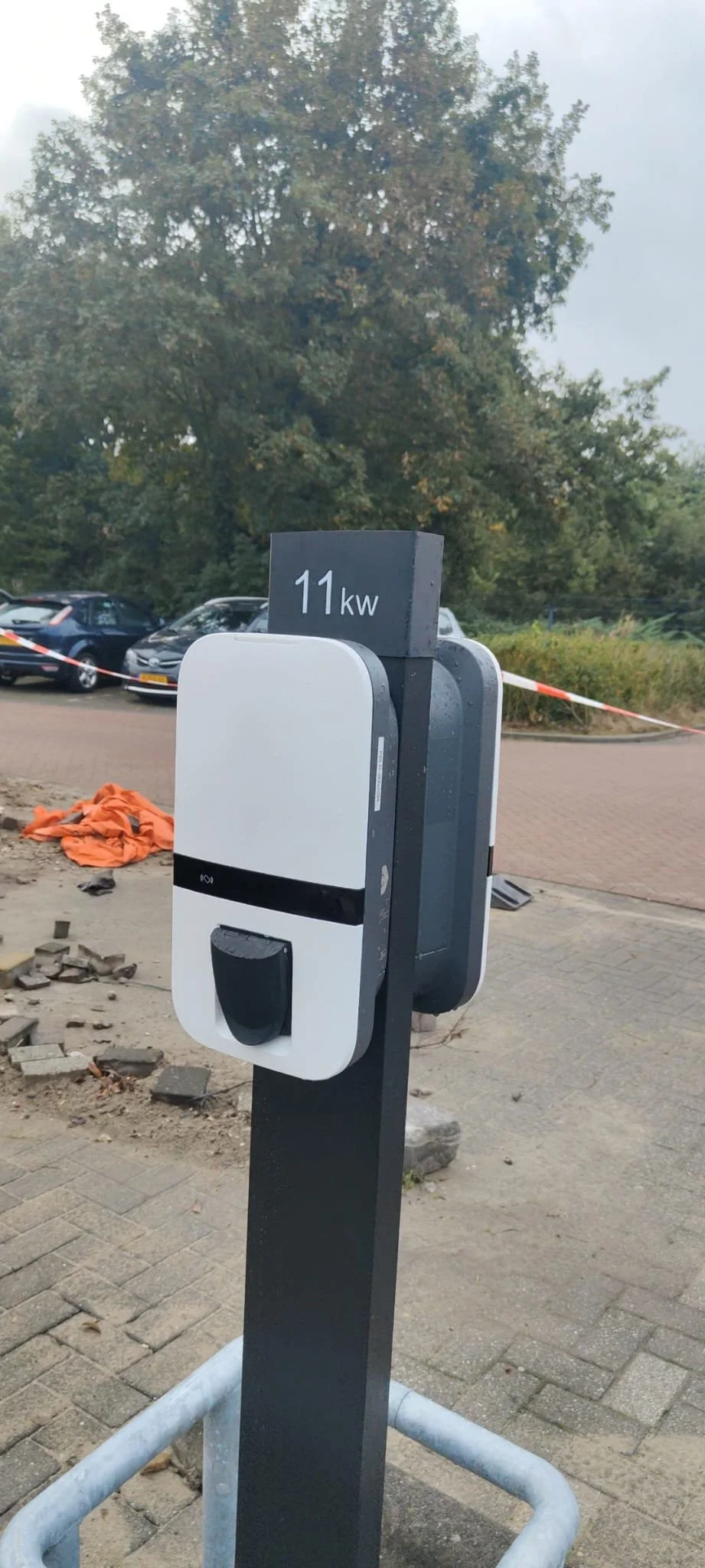 Oplaadpaal voor elektrische auto's, met een bordje erop dat aangeeft dat het 11 kilowatt levert, geplaatst naast een parkeerplaats en een groen landschap op de achtergrond. Bordje is 3D geprint.