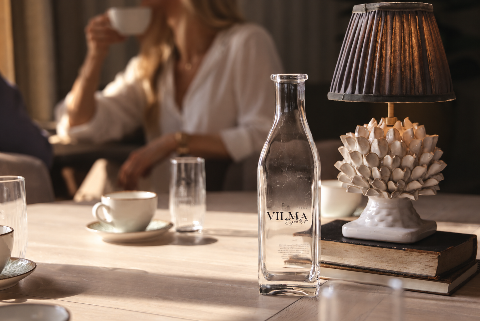 Et bord med karaffel, kaffekopp, glass og et lampe oppå noen bøker. I bakgrunnen sitter det en dame og drikker kaffe.