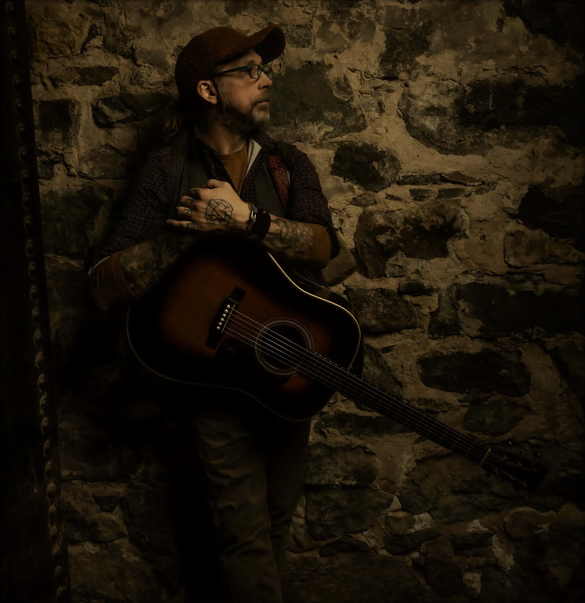 A man performing live music by singing and playing an acoustic guitar in a cozy, warmly lit room with orange wallpaper, framed posters on the wall, a fireplace, a table lamp, and various boxes and equipment around.