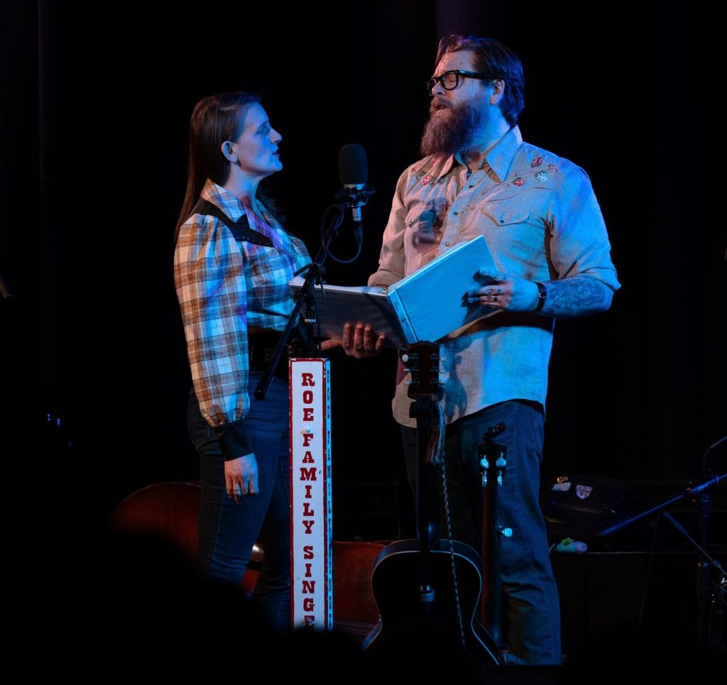 A man performing live music by singing and playing an acoustic guitar in a cozy, warmly lit room with orange wallpaper, framed posters on the wall, a fireplace, a table lamp, and various boxes and equipment around.