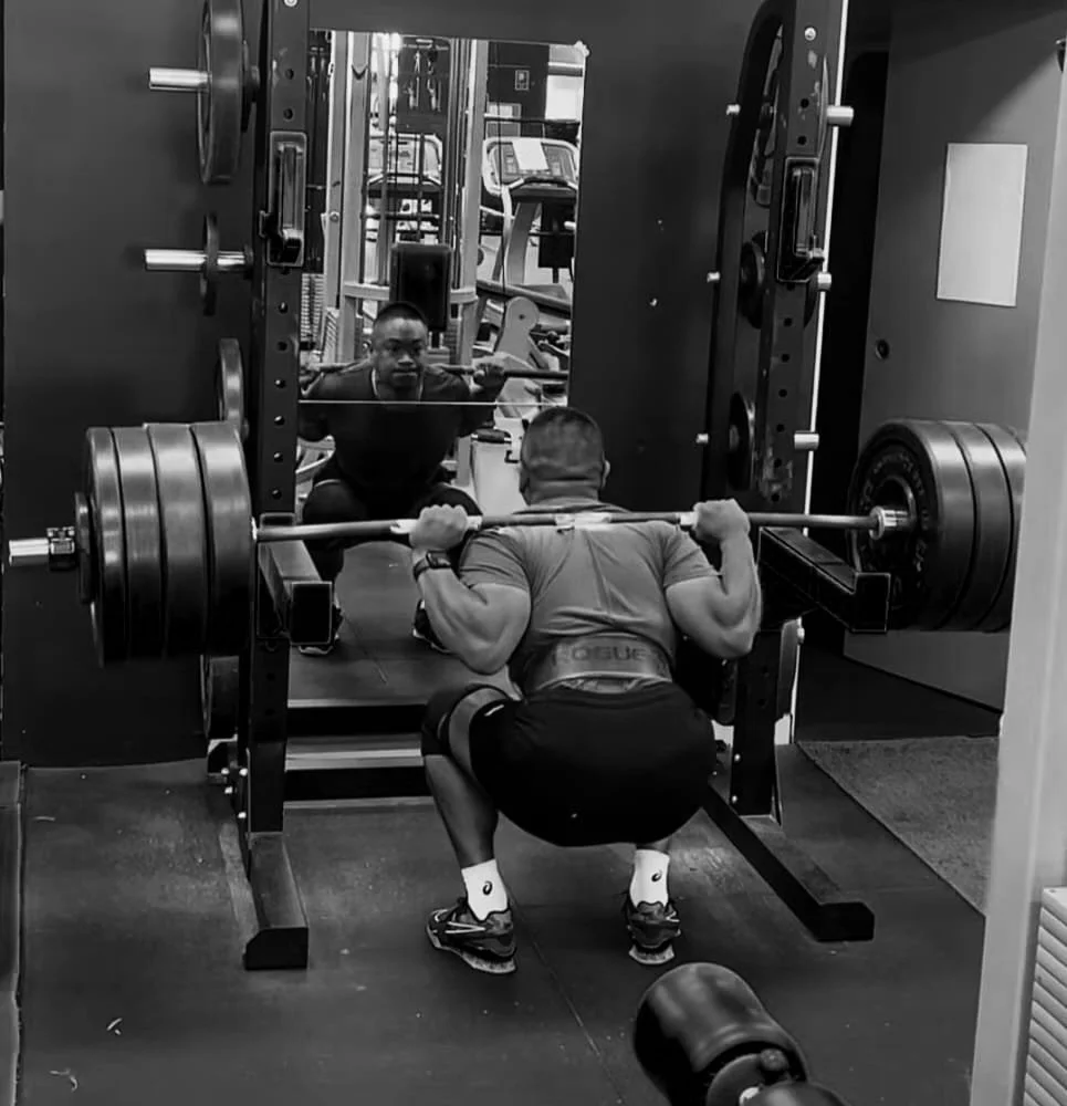 A man squatting and lifting a loaded barbell in a gym, with his reflection visible in a mirror behind him.