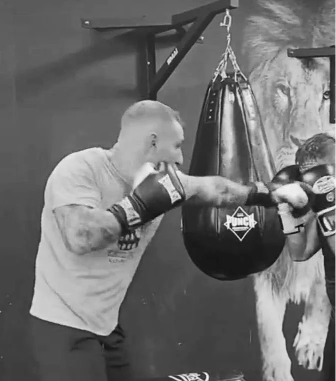 Two fighters sparring with boxing gloves in a gym, punching a black punching bag hanging from the ceiling.