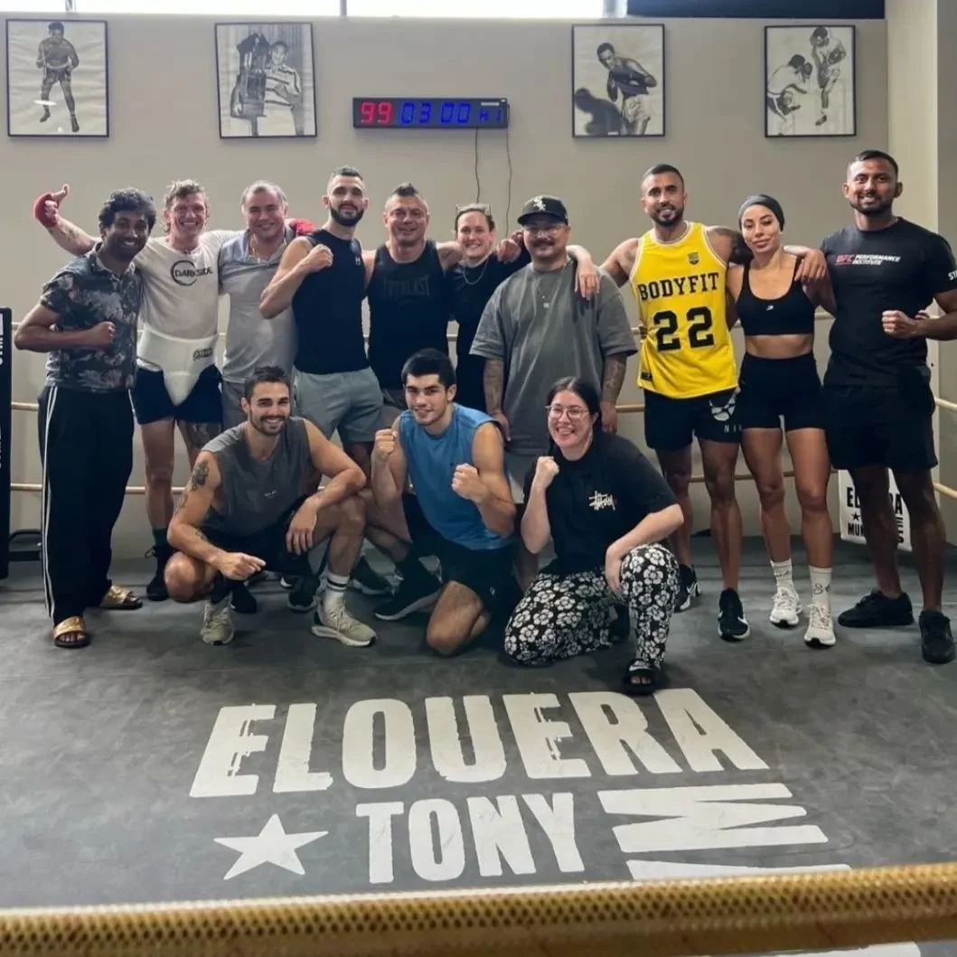 A group of people in athletic wear posing together in a gym with a boxing ring. The floor has the words 'ELOUER TONY'. There are framed photos on the wall and a digital clock showing 3:00.
