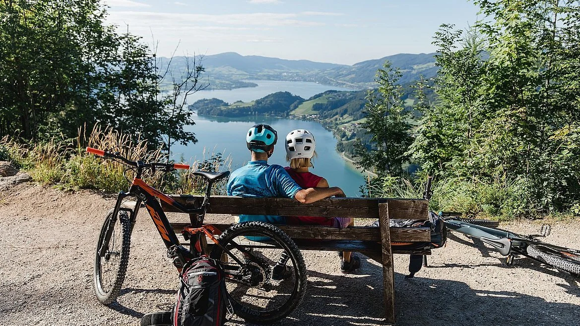 Zwei Personen mit Fahrradhelmen sitzen auf einer Bank mit Blick auf einen See und Berge im Hintergrund, umgeben von Bäumen und Fahrrädern.