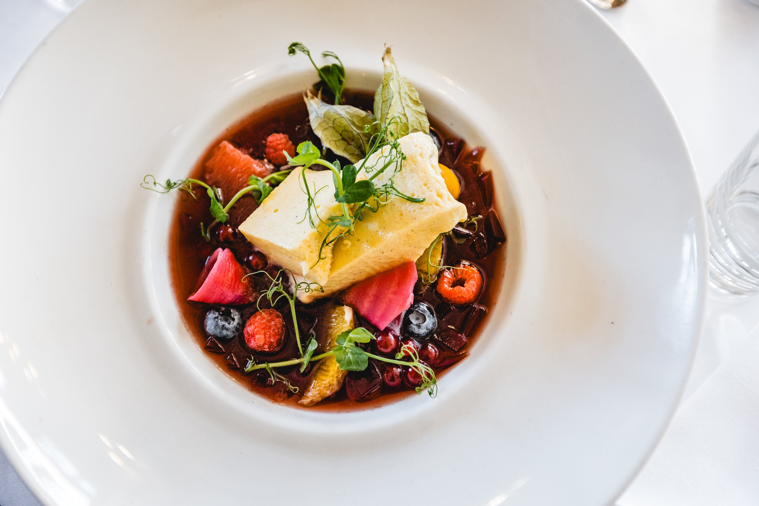 Ein Dessert in einem weißen Teller mit einer Creme oder Panna Cotta auf einem roten Fruchtkompott, garniert mit Beeren, Kräutern und essbaren Blütenblättern.