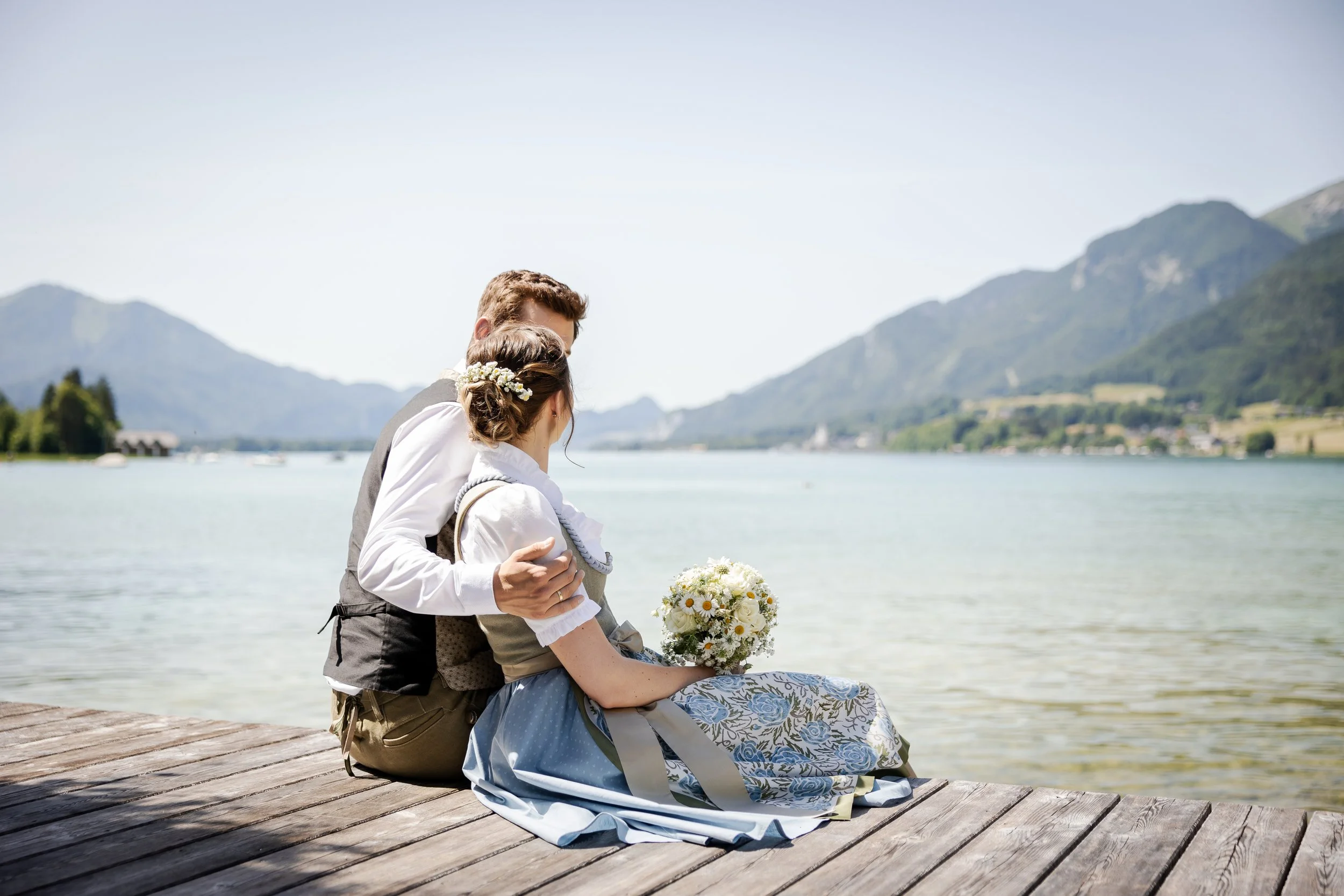 Paar in traditioneller bayerischer Tracht sitzt auf einem Steg am See mit Bergen im Hintergrund.