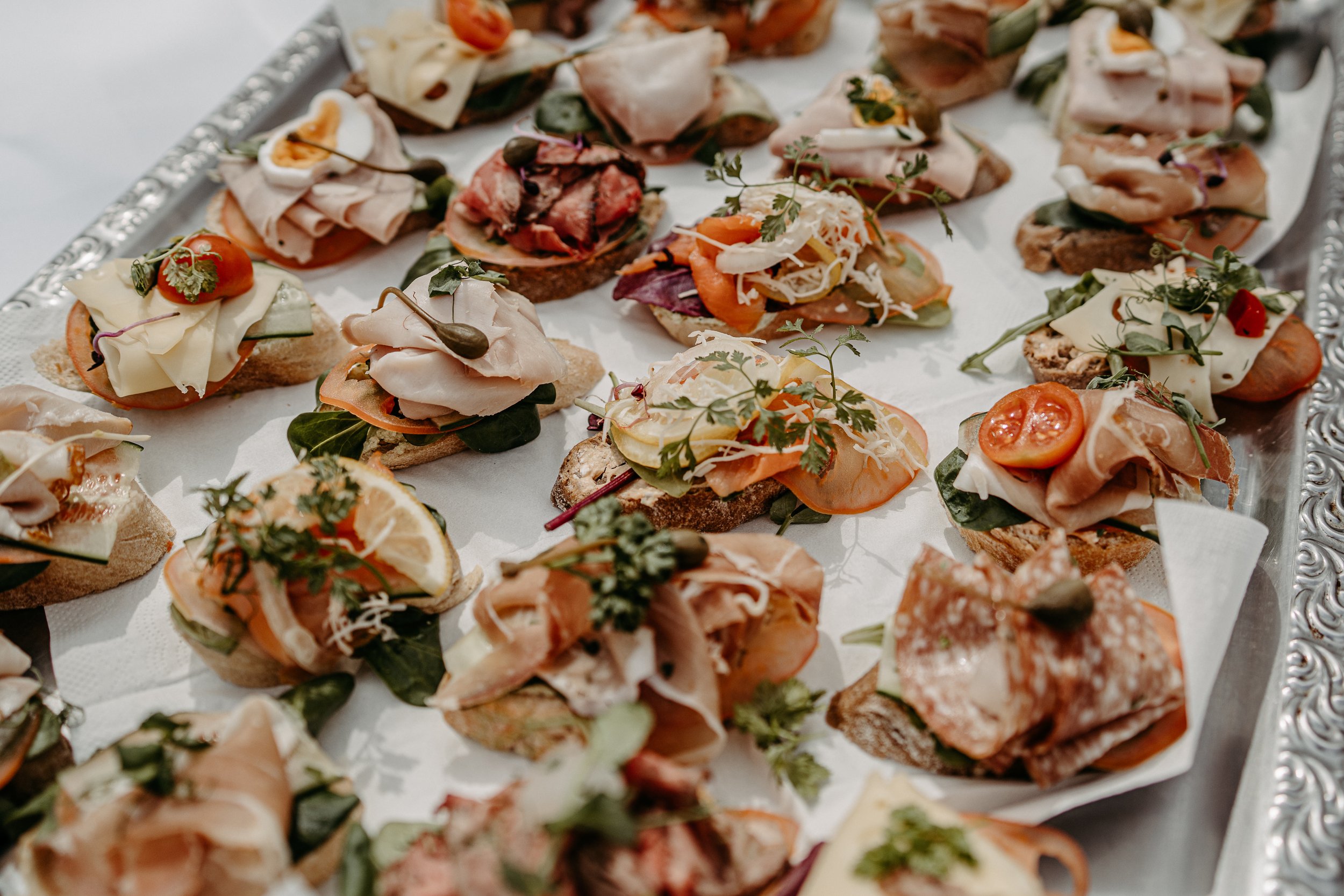 Eine Vielzahl von belegten Brötchen auf einem Tablett, garniert mit verschiedenen Zutaten wie Schinken, Käse, Tomaten, Kräutern und Gemüse.