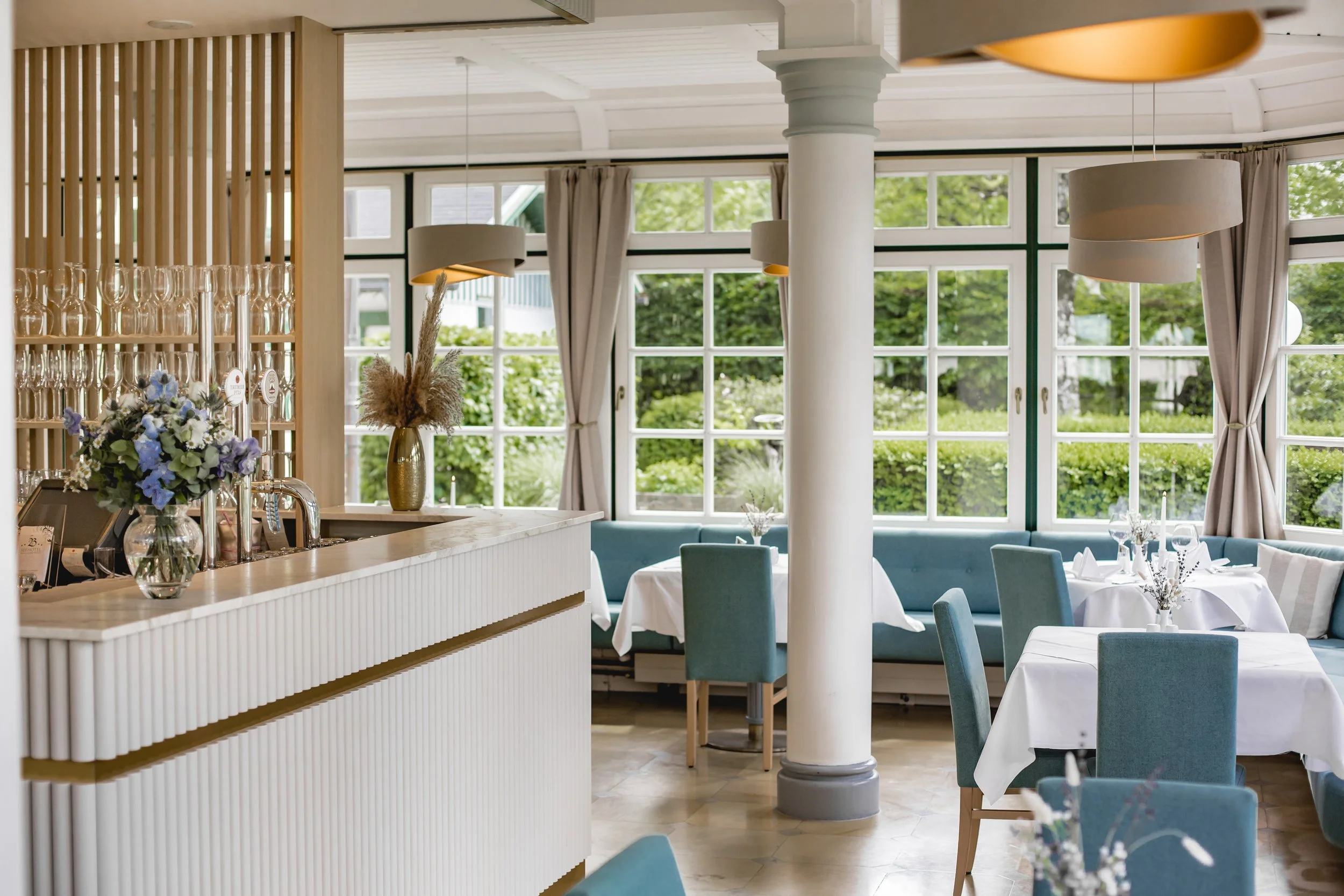 Ein elegantes Restaurant mit blauen Stühlen und weißen Tischdecken. Große Fenster lassen viel Licht herein. Eine Theke mit Blumenarrangement ist sichtbar. Die Einrichtung wirkt modern und einladend.