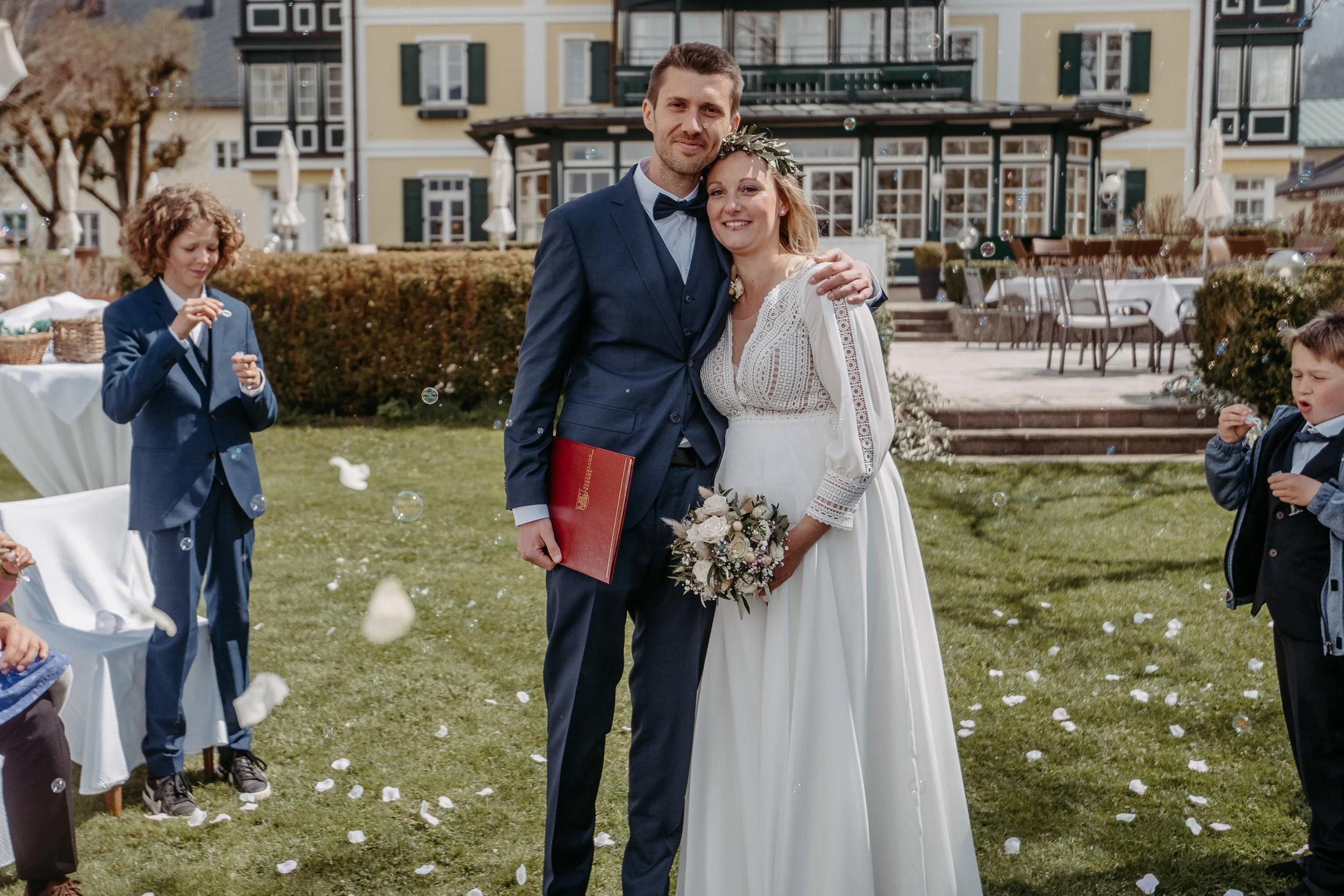 Hochzeit, glückliches Paar in Hochzeitskleidung draußen, umgeben von Kindern, die Seifenblasen blasen, im Hintergrund ein Gebäude.