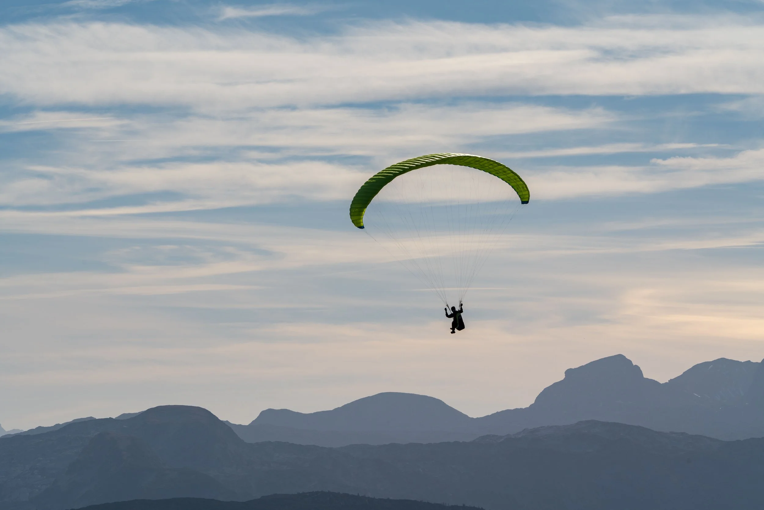 Person beim Gleitschirmfliegen vor Bergkulisse im blauen Himmel mit Wolken.