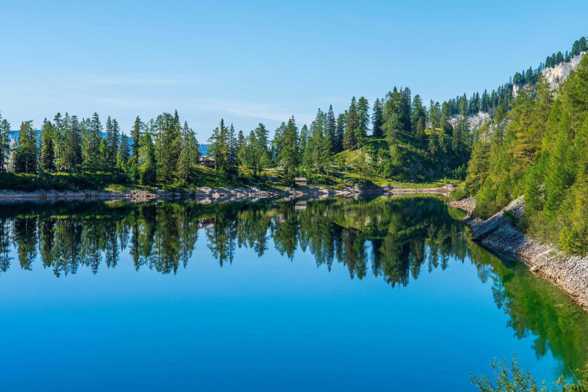 Ein klarer See mit Spiegelung von Bäumen und Bergen im Hintergrund unter blauem Himmel.