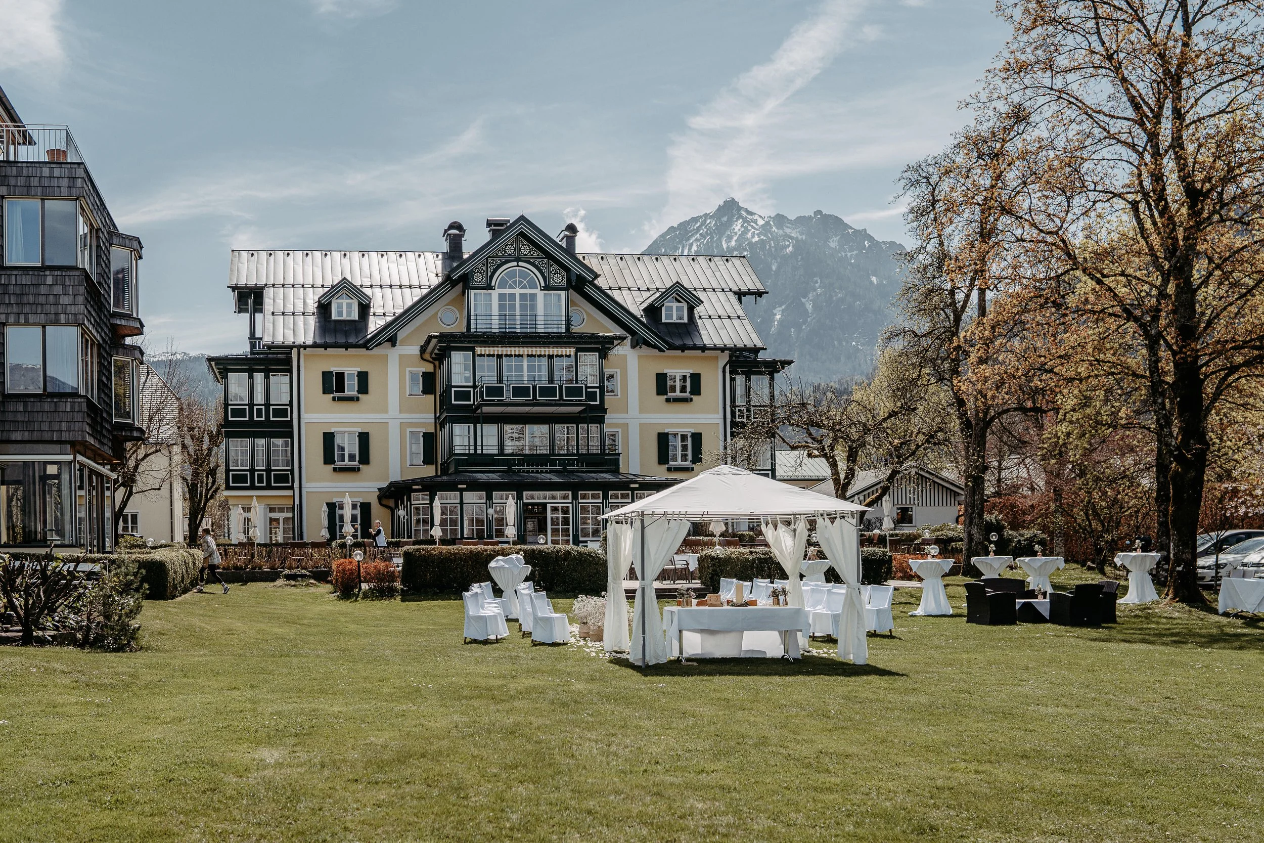 Historisches Gebäude vor Bergen, Rasenfläche mit Pavillon und Tischen, umgeben von Bäumen.