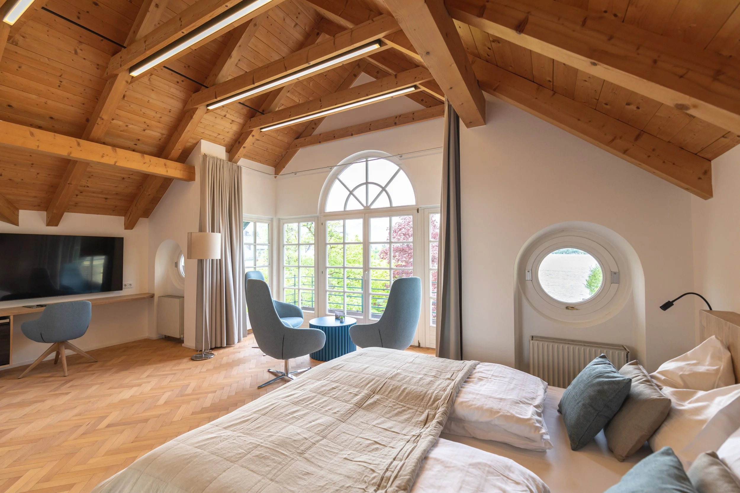 Geräumiges Schlafzimmer mit Holzdecke, großem Fenster, Balkonblick und modernen Möbeln, darunter ein Bett, Fernseher und zwei blauen Sesseln.