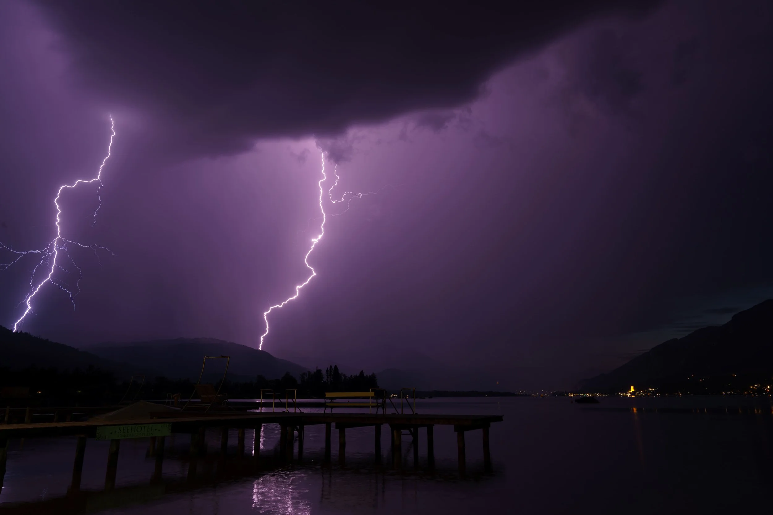 Blitzeinschläge bei Nacht über einem dunklen See mit einem Steg im Vordergrund und Lichtern in der Ferne.