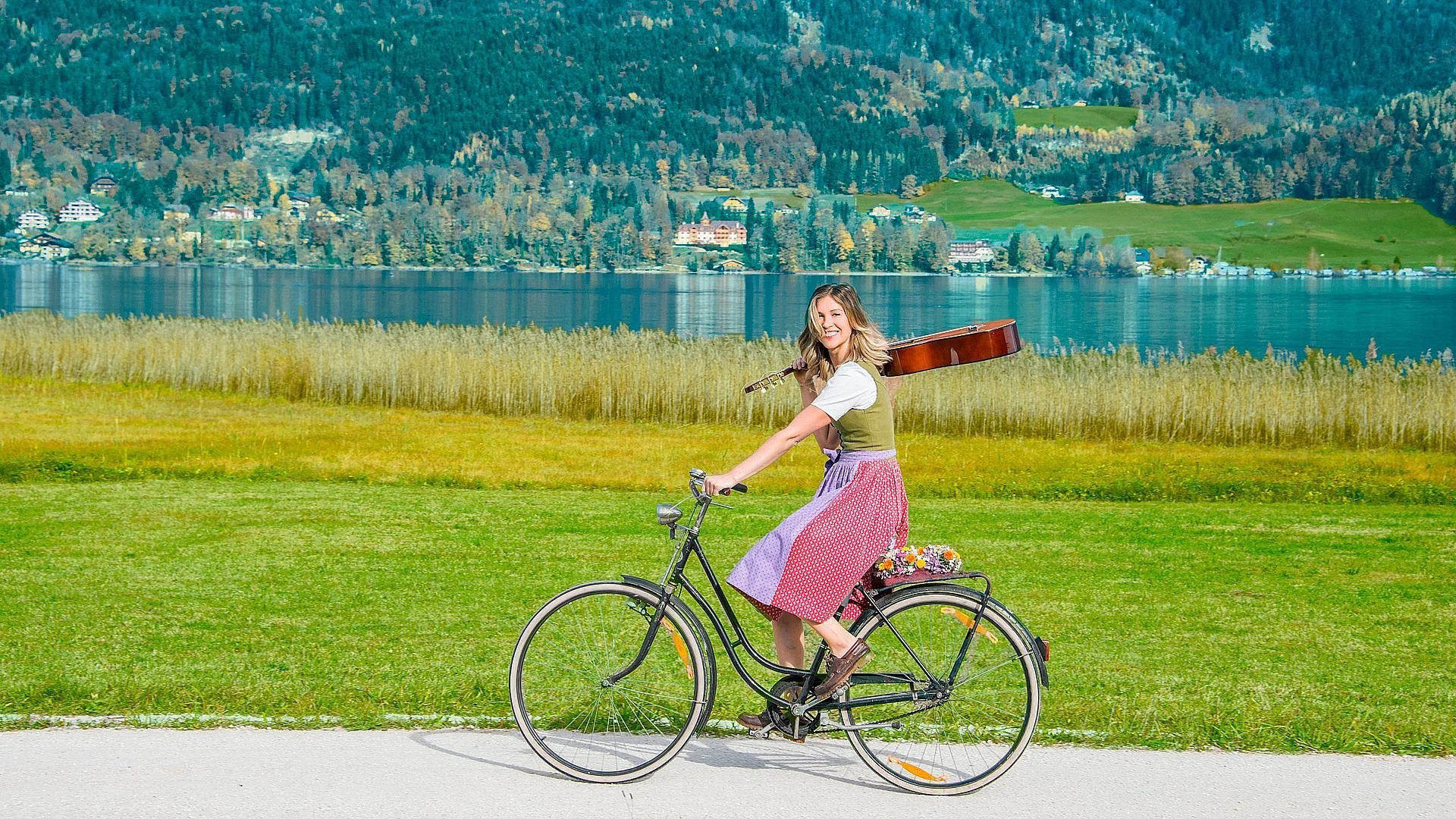 Eine Frau in traditioneller Kleidung fährt auf einem Fahrrad mit einer Gitarre auf dem Rücken an einem See entlang.
