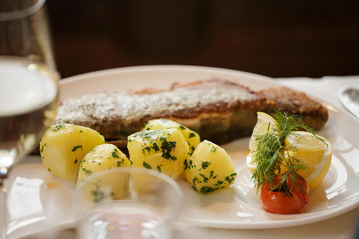 Ein Teller mit gebratenem Fisch, Petersilienkartoffeln, Zitronenspalten, Dill und einer Kirschtomate.