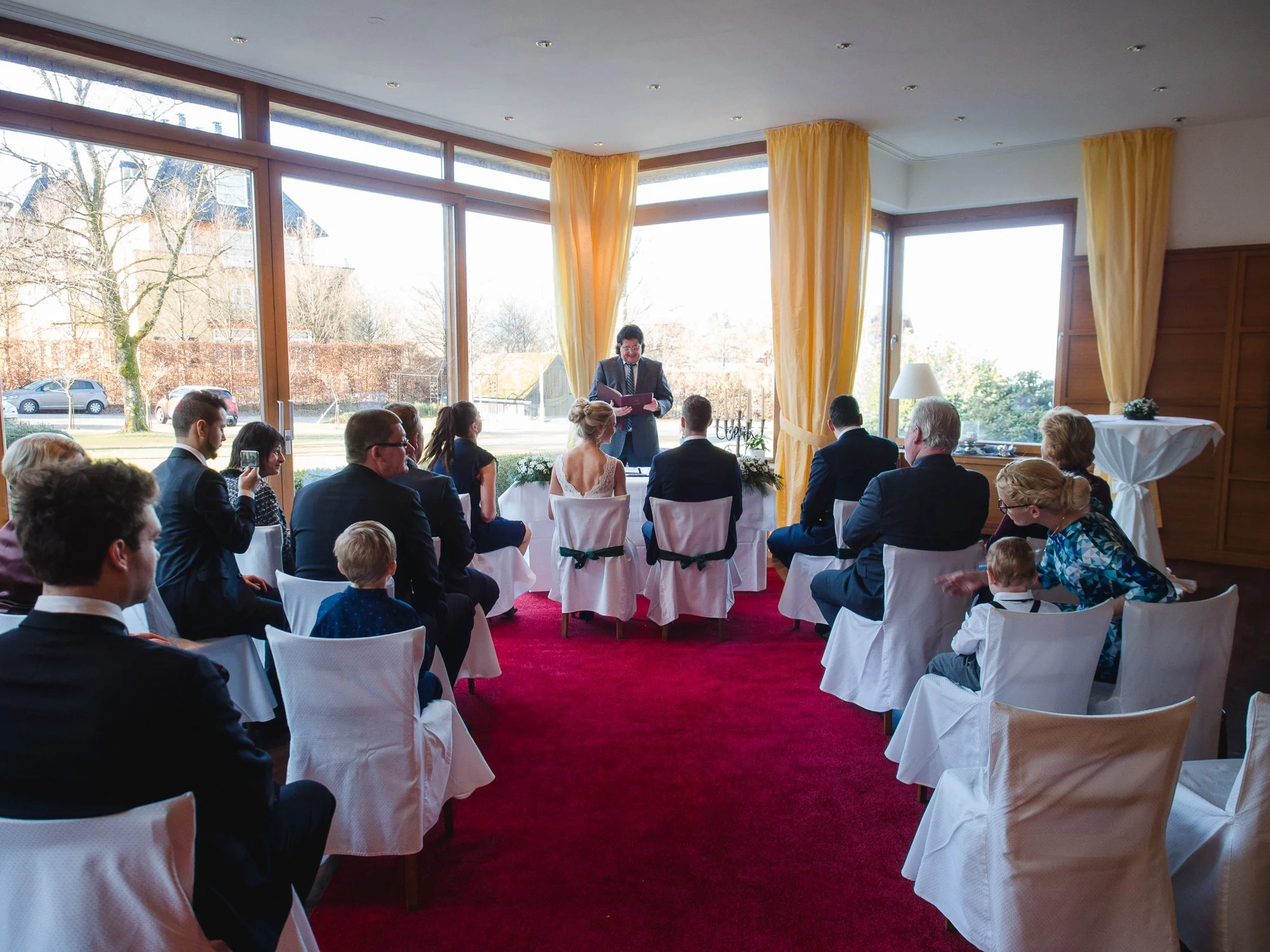 Eine kleine Versammlung in einem Raum mit großen Fenstern und gelben Vorhängen. Menschen sitzen auf weißen Stühlen mit Hussen, während ein Redner vorne steht. Einige tragen elegante Kleidung. Draußen sind Bäume und ein geparktes Auto sichtbar.