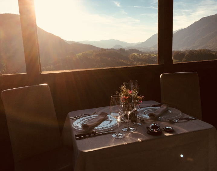 Ein Esstisch mit zwei eingedeckten Plätzen, Gläsern und einem Blumenstrauß, in einem Raum mit Blick auf eine Berglandschaft bei Sonnenuntergang.