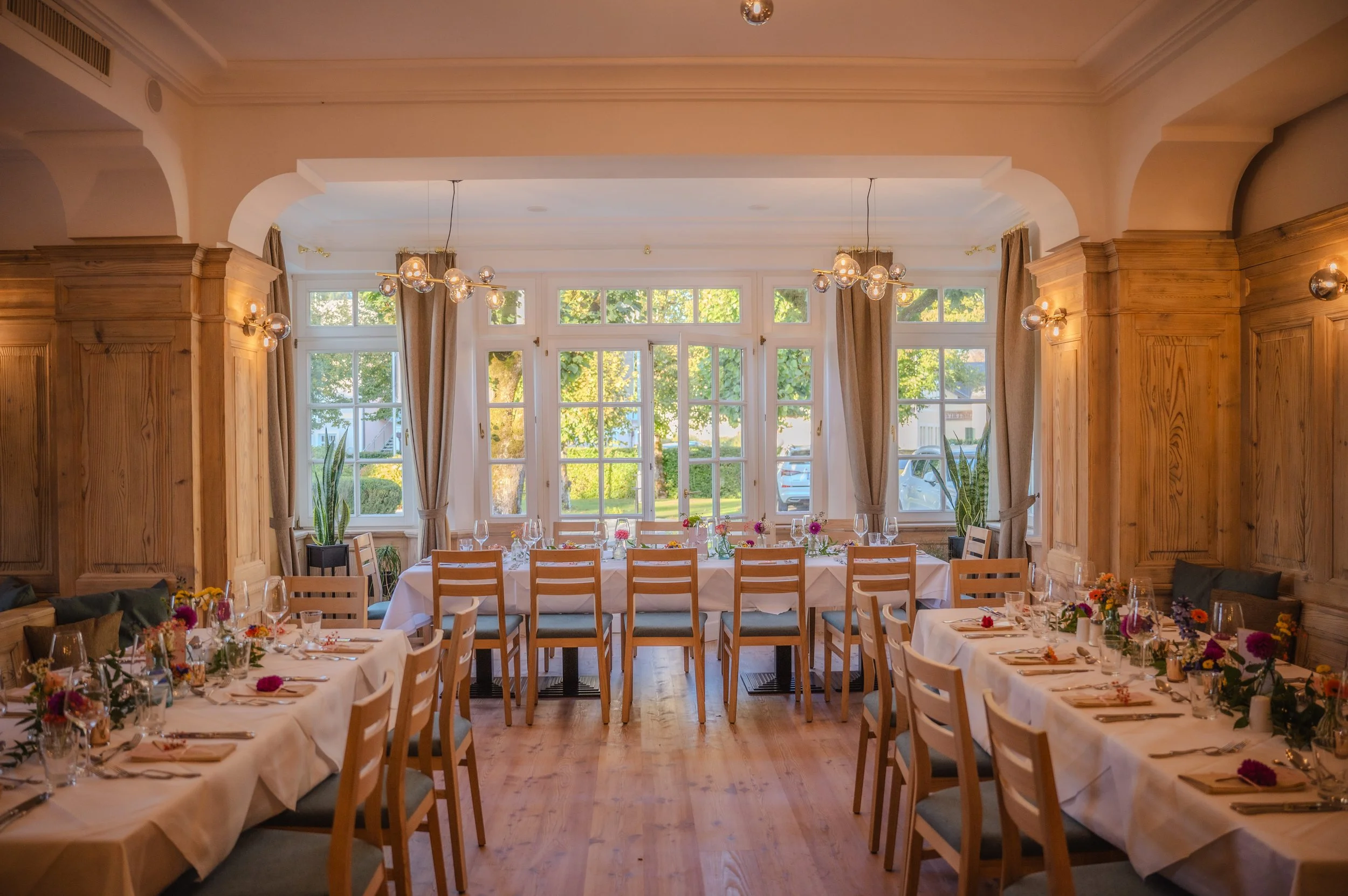 Eleganter Speisesaal mit gedeckten Tischen, Holzstühlen und großen Fenstern. Die Tischdekoration umfasst Blumen und Kerzen. Es herrscht eine warme und einladende Atmosphäre.