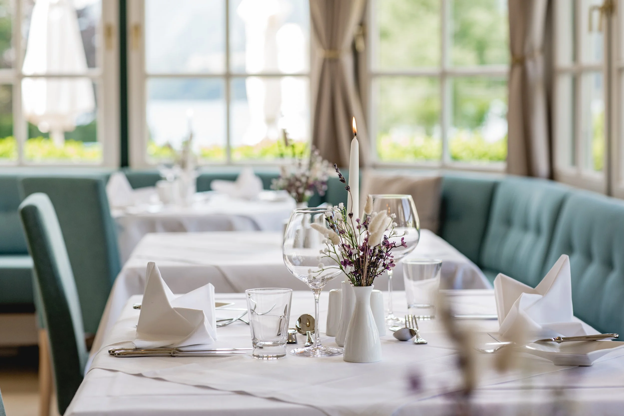 Elegantes Restaurant mit weiß gedeckten Tischen, Glasvasen mit Blumen, gefalteten Servietten und brennender Kerze, große Fenster im Hintergrund.