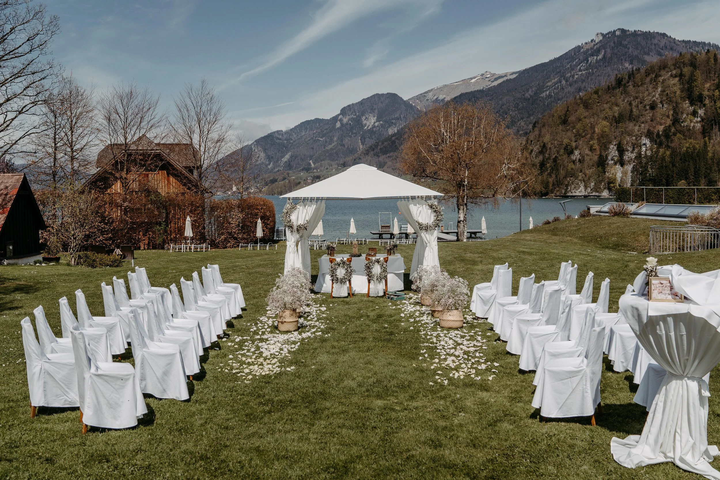 Freiluft-Hochzeitszeremonie am See, mit weißen Stühlen und Blütenblättern, Bergkulisse im Hintergrund.