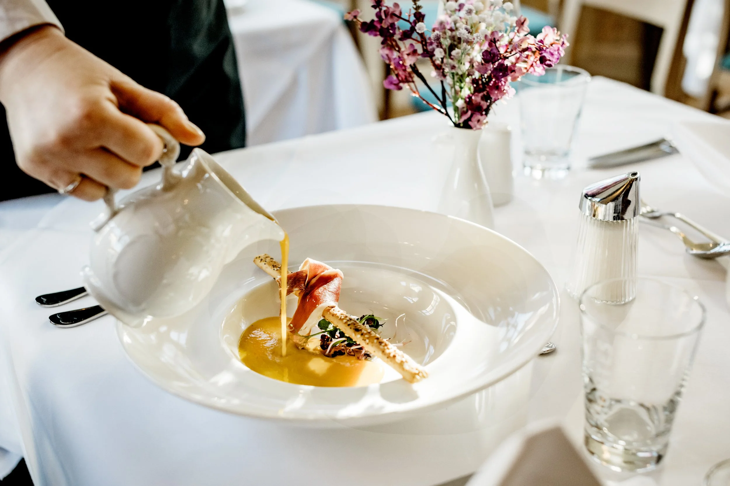 Gourmet-Suppe in einem tiefen Teller, dekoriert mit Brotstangen und Kräutern; eine Hand gießt Suppe aus einem Krug. Im Hintergrund sind Blumen und Tischaccessoires sichtbar.