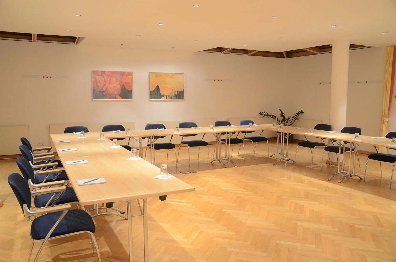 Konferenzraum mit Holzboden, U-förmiger Tischanordnung, blau gepolsterten Stühlen, Notizblöcken, Stiften und Wassergläsern, helle Wände mit zwei Gemälden und Zimmerpflanze.