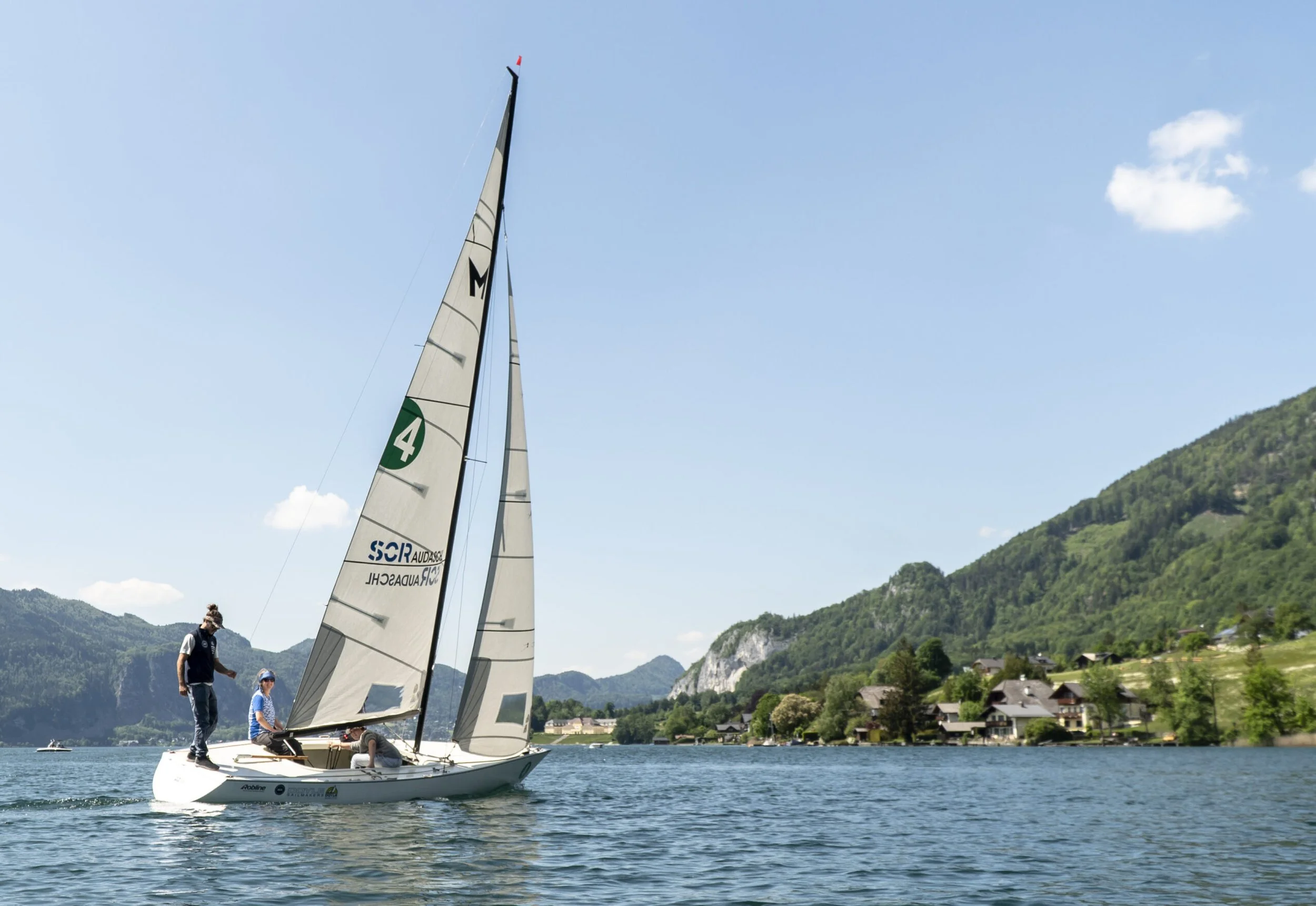 Segelboot mit drei Personen auf einem See, umgeben von Bergen und Häusern im Hintergrund bei klarem Himmel.