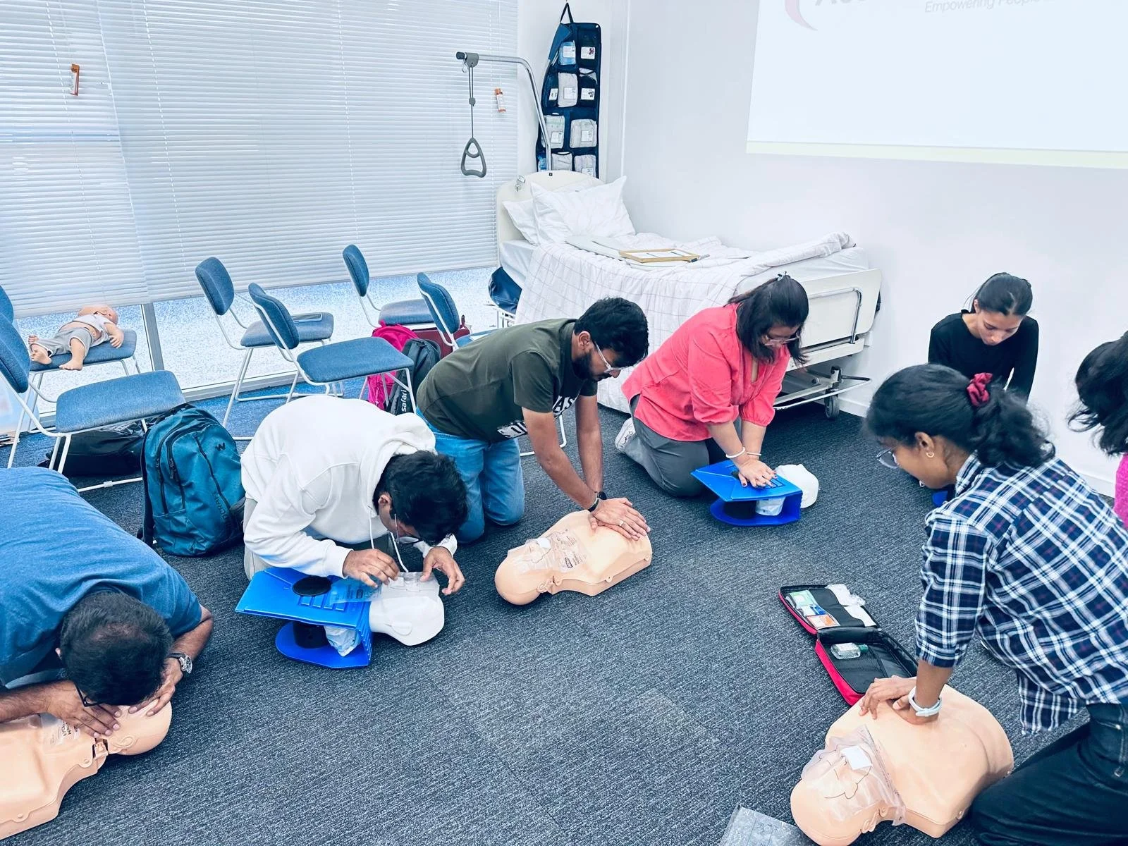 Group of people practicing CPR on mannequins in a classroom setting, with chairs, a hospital bed, and medical supplies visible in the background.