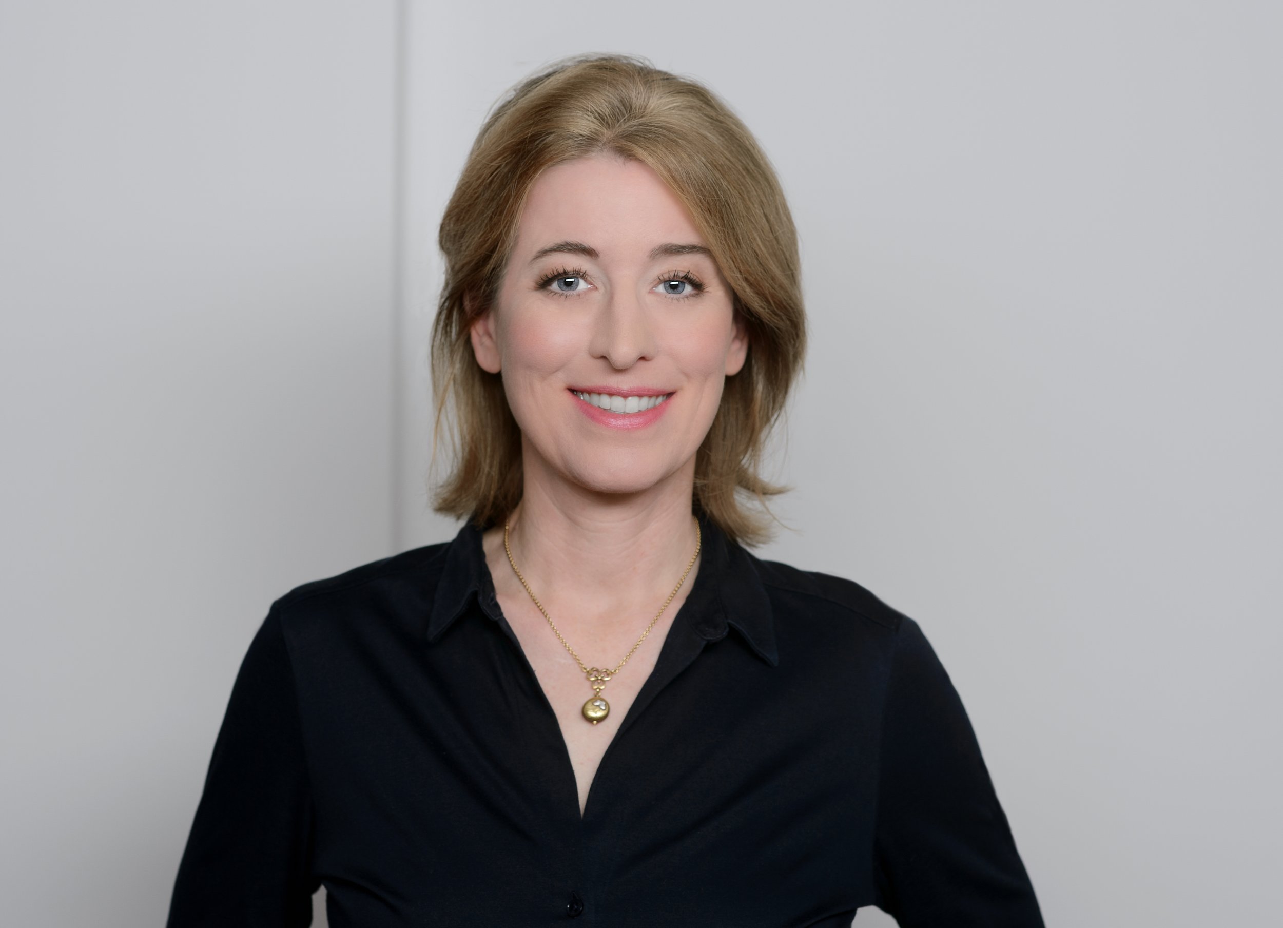 Ein Porträt einer lächelnden Frau mit mittellangen, blonden Haaren, trägt eine schwarze Bluse und eine goldene Halskette vor einem neutralen Hintergrund.