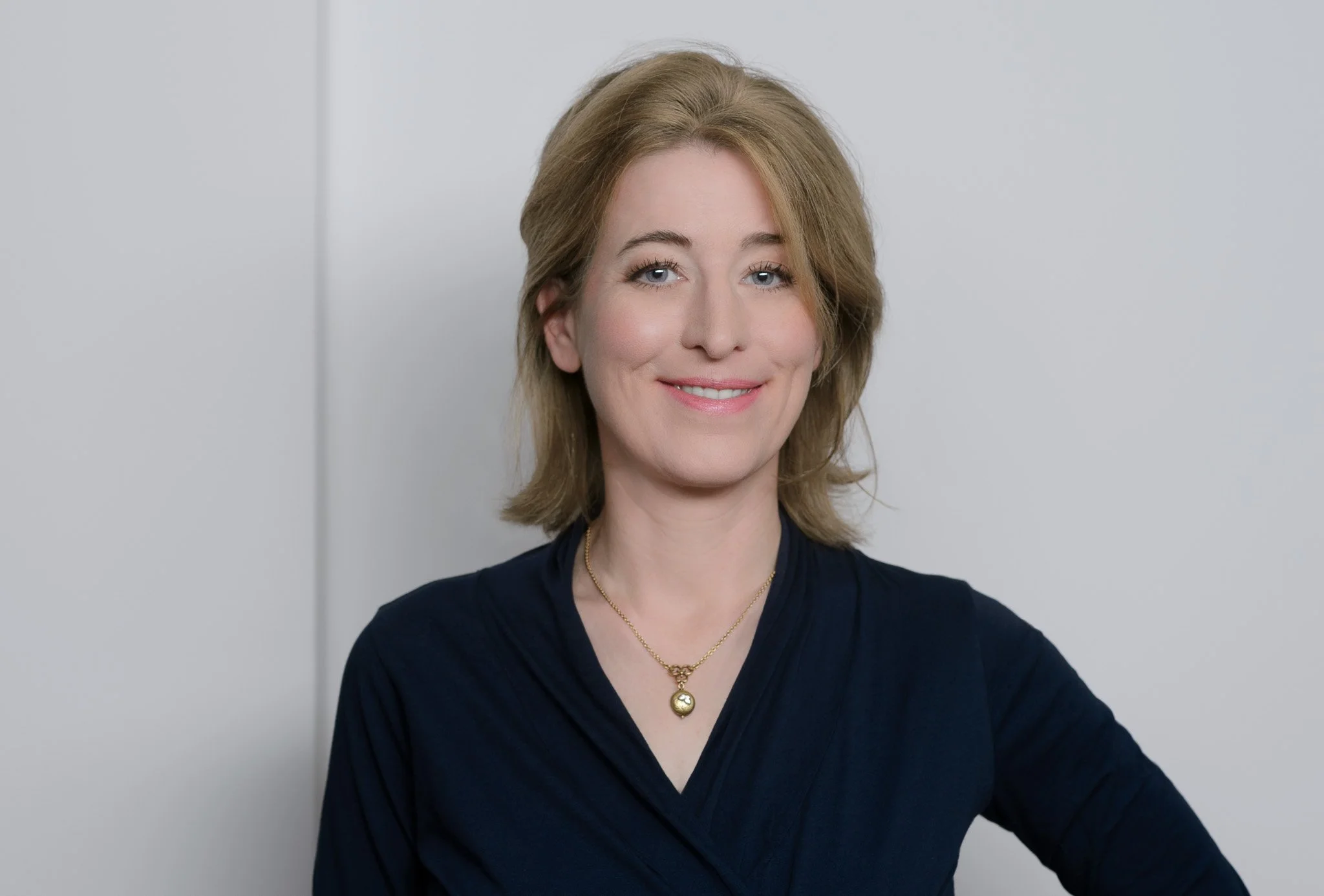 Eine lächelnde Frau mit mittellangem, blondem Haar trägt eine dunkle Bluse und eine goldene Halskette, steht vor einer hellgrauen Wand.