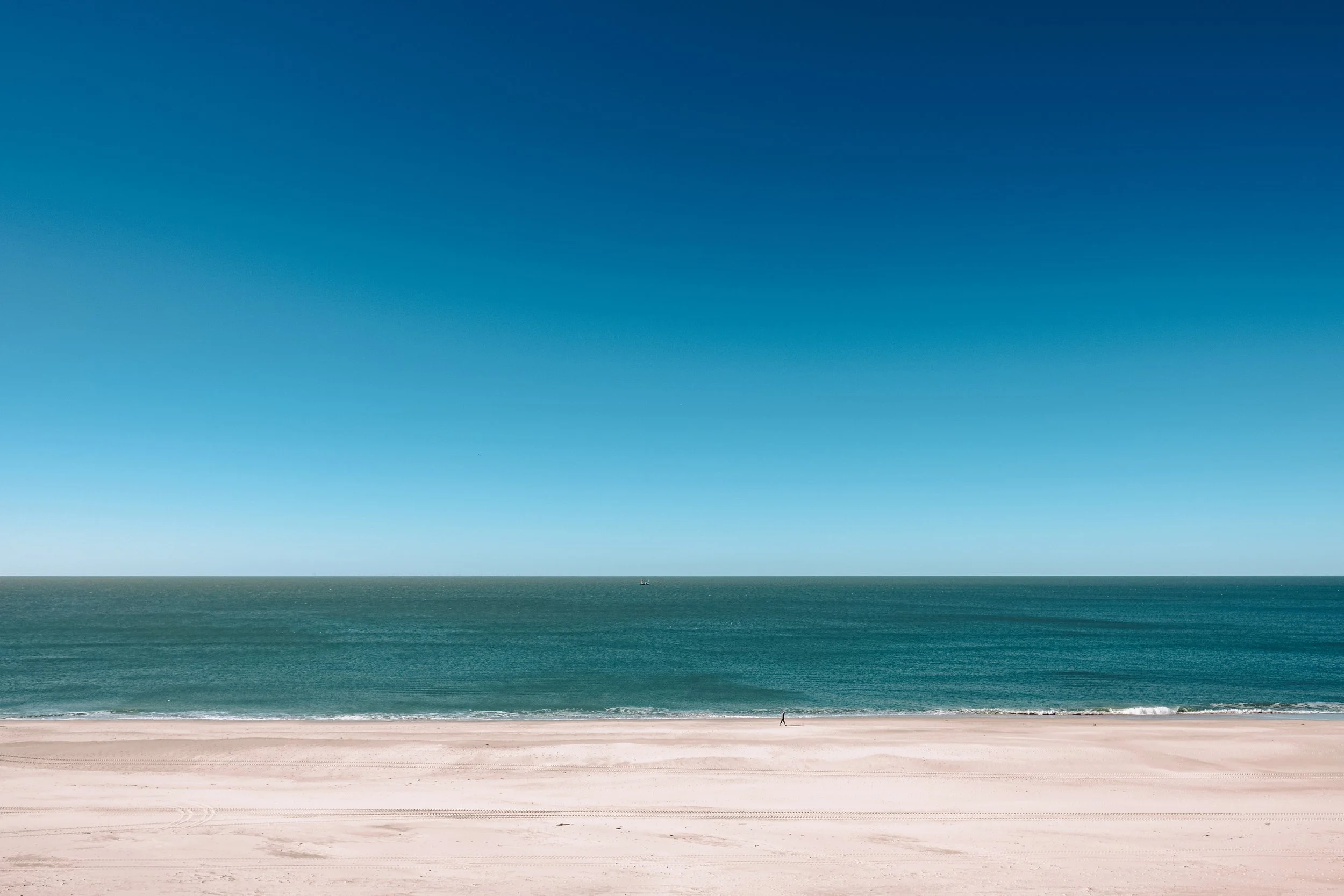 Leerer Strand mit Sand und Wasser, im Hintergrund das Meer und ein klarer blauer Himmel.