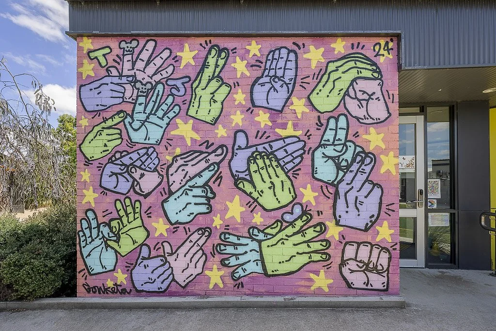 Colourful mural on a wall featuring multiple stylised hand gestures in pink, green, and blue tones, surrounded by yellow stars, set against a pink background.