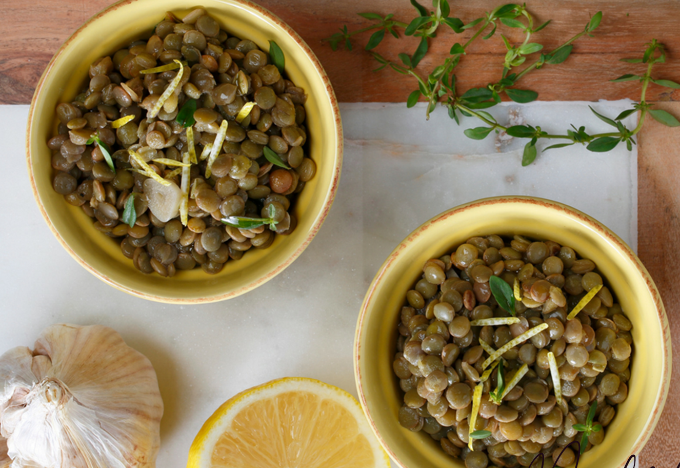 lemony lentils in two yellow bowls on a wooden table with garlic, lemon, and herbs arranged nearby