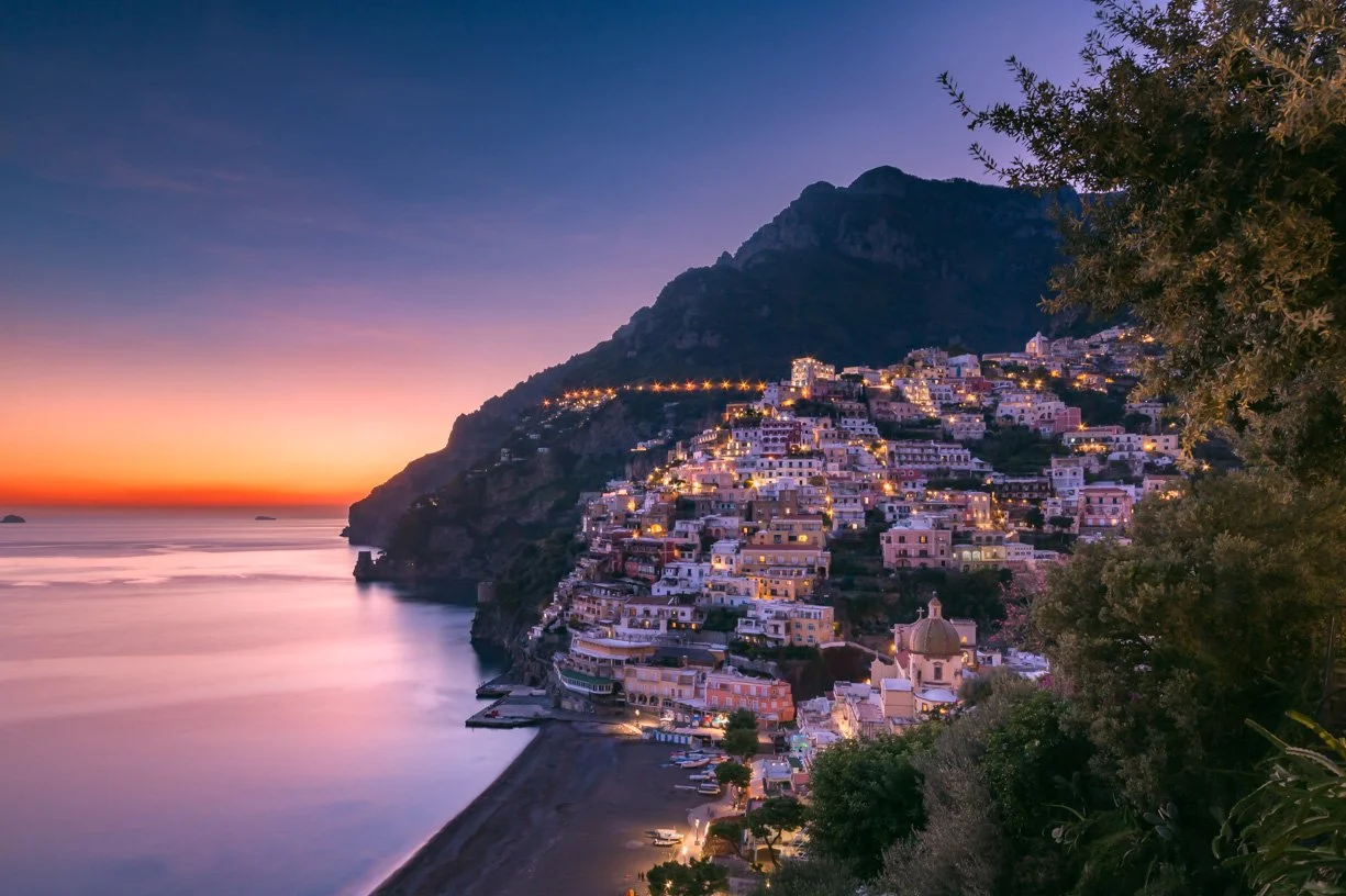 Coastal village at sunset with hillside buildings, trees in the foreground, and calm ocean waters.