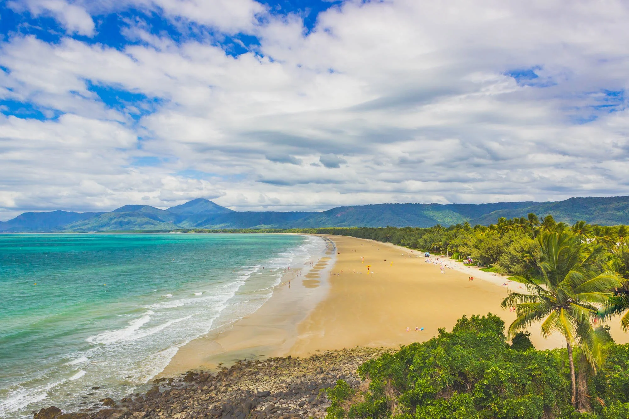 A tropical beach with turquoise waters, a sandy shoreline, lush green palm trees, mountains in the background, and a partly cloudy sky.