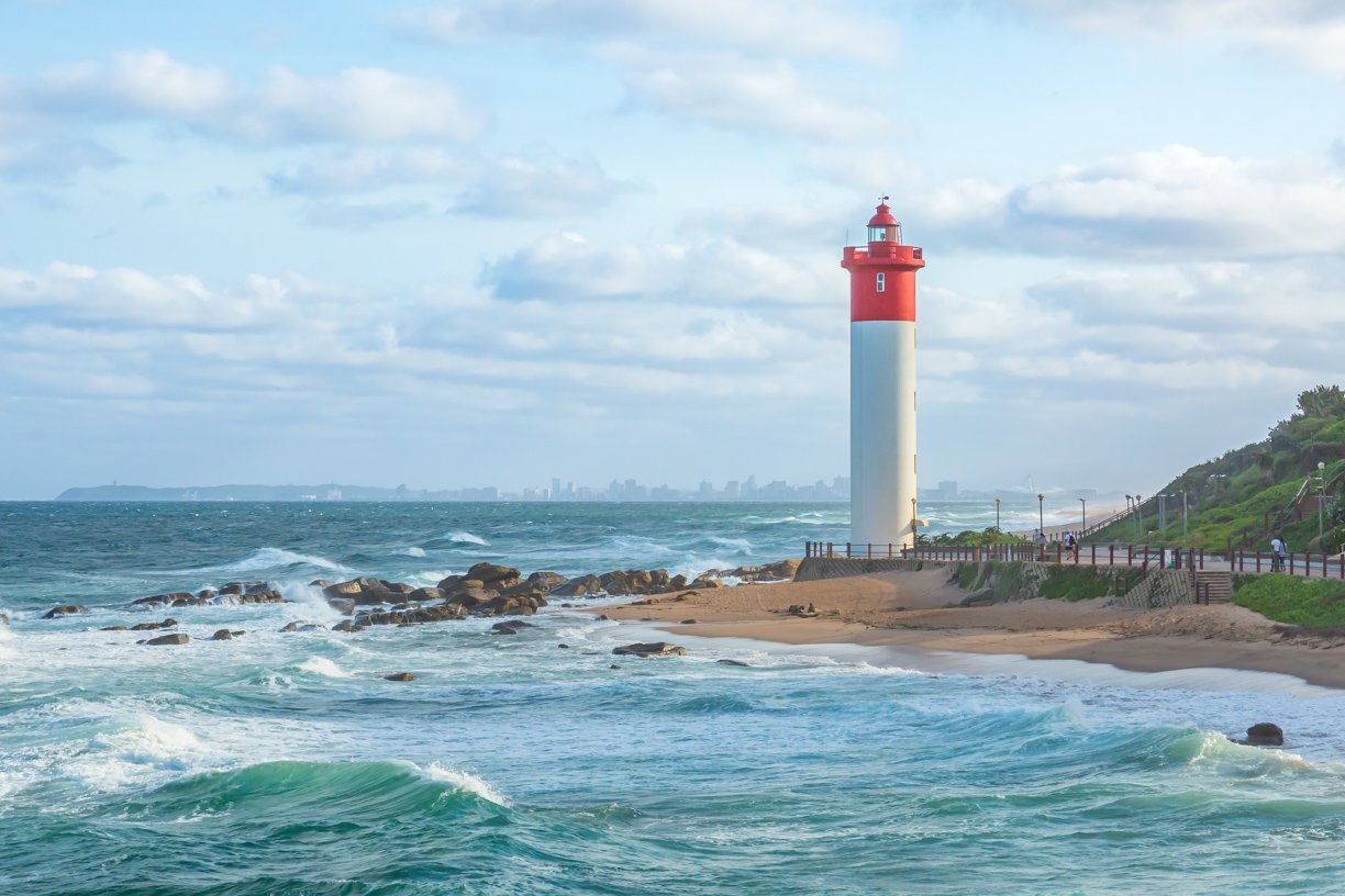 A coastal scene featuring a red and white lighthouse on the sandy beach. The ocean waves crash against rocks, with a city skyline visible in the distance under a partly cloudy sky. A walkway runs along the shoreline with a few people walking.