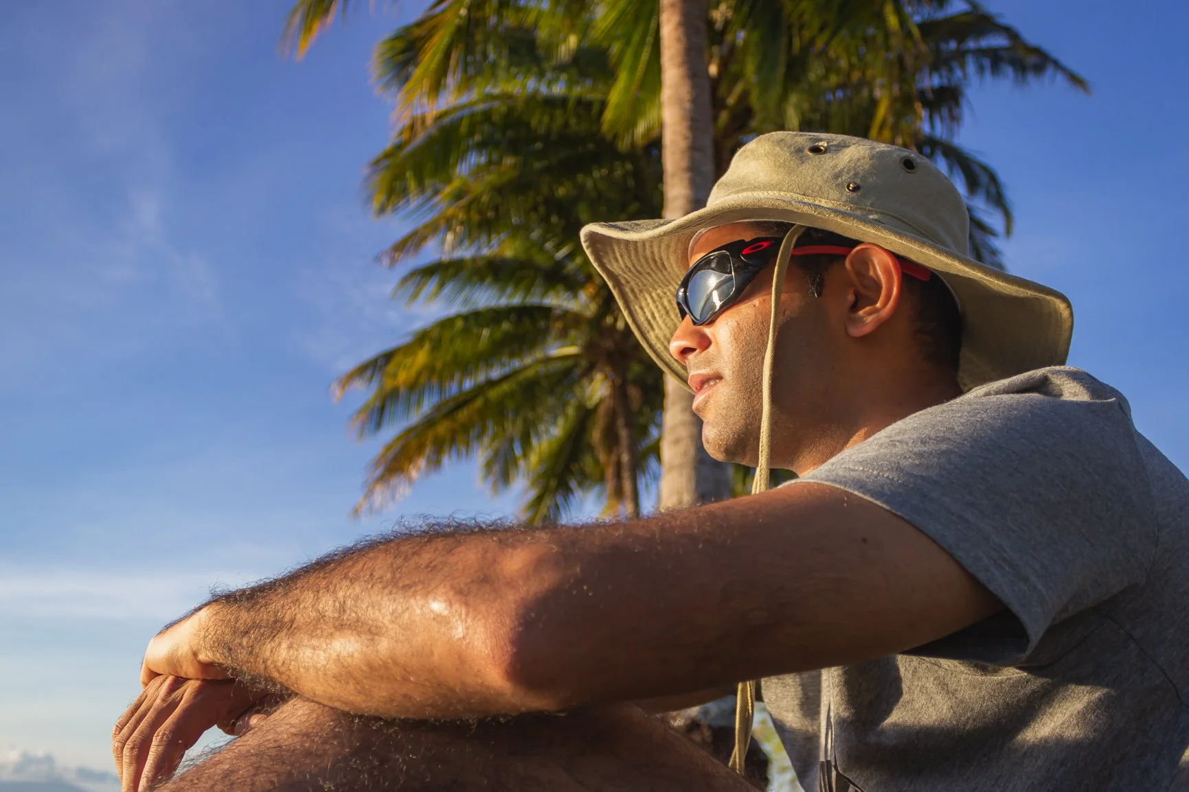 A man wearing sunglasses, a wide-brimmed hat, and a gray t-shirt sits outdoors with palm trees and a blue sky in the background.