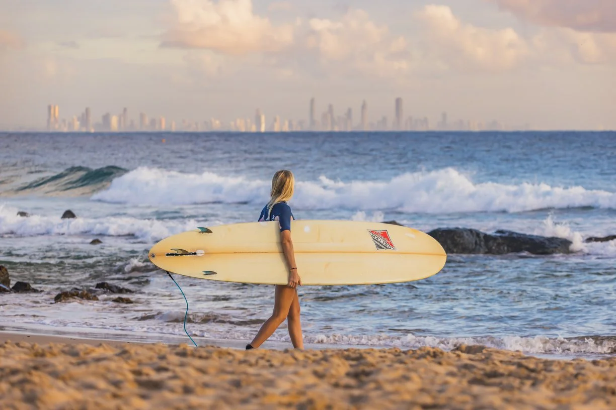 A woman walking on the beach with a surfboard under her arm, looking out at the ocean with a city skyline in the distance.