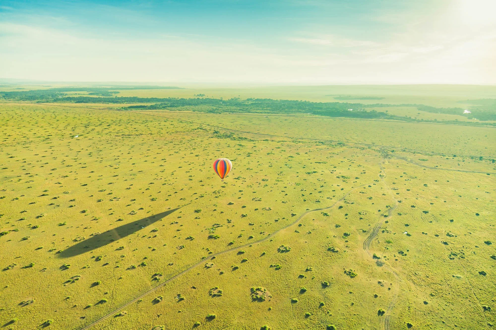 A hot air balloon with bright orange, yellow, and blue vertical stripes floating over vast stretch of African savannah landscape.