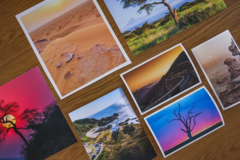 A collection of eight landscape photographs spread out on a wooden surface, featuring deserts, forests, coastlines, mountains, a road, a sunset, and a leafless tree.