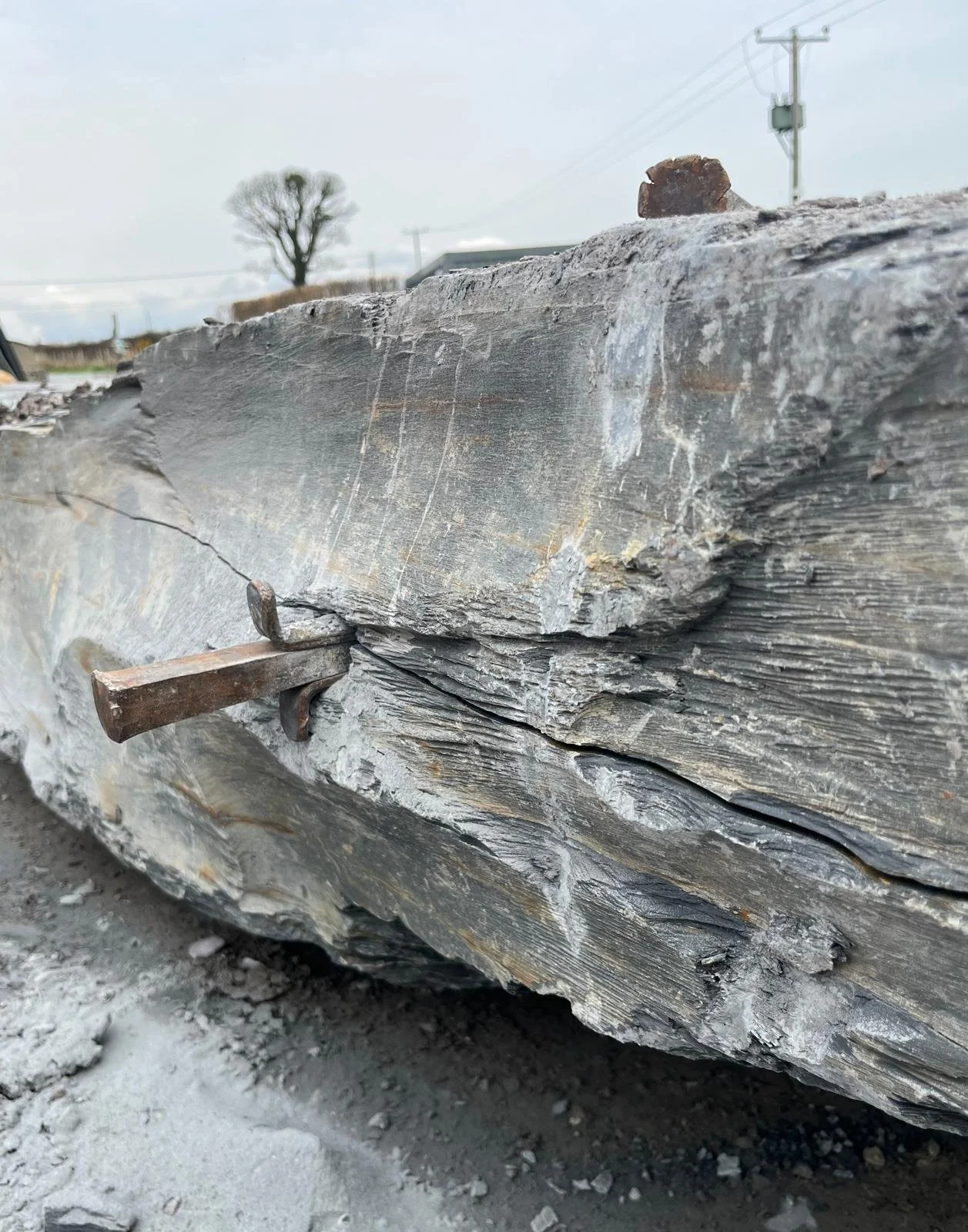 Step by step view of the plug and feather method: two curved metal shims (feathers) and a central wedge (plug) inserted into drilled holes in stone, then hammered to create controlled cracks and split the slate.