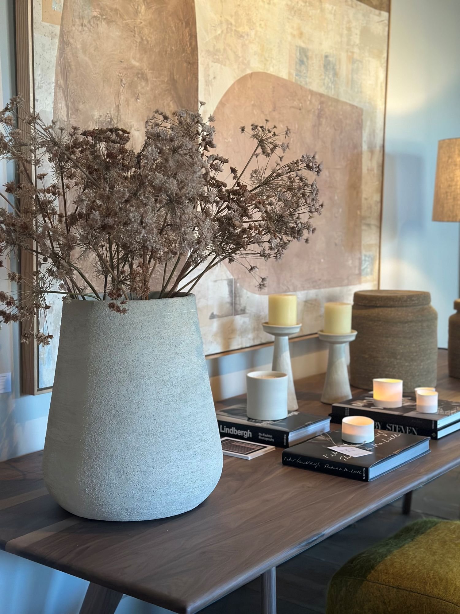 A large textured white vase with dried flowers is on a wooden table. Behind it, a beige abstract artwork, three white candles on small stands, and a stack of fashion books are visible, along with a table lamp and a mustard-colored chair.