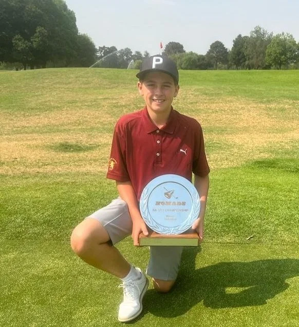Young boy kneeling on the grass holding a trophy, outdoors on a golf course. He is wearing a black cap with a white 'P' and a red polo shirt. The background shows trees and a golf flag.