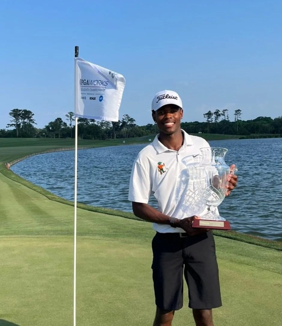 A golfer standing on a golf course near a water hazard, holding a trophy, with a flag marking the hole in the background under a clear blue sky.