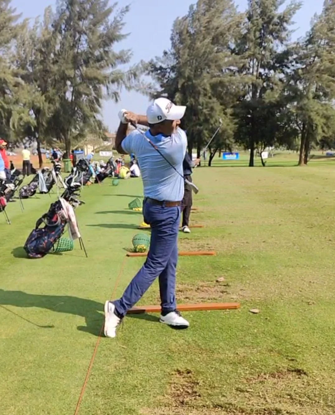 A golfer in gray polo and navy pants swinging a golf club on a driving range with trees in the background.