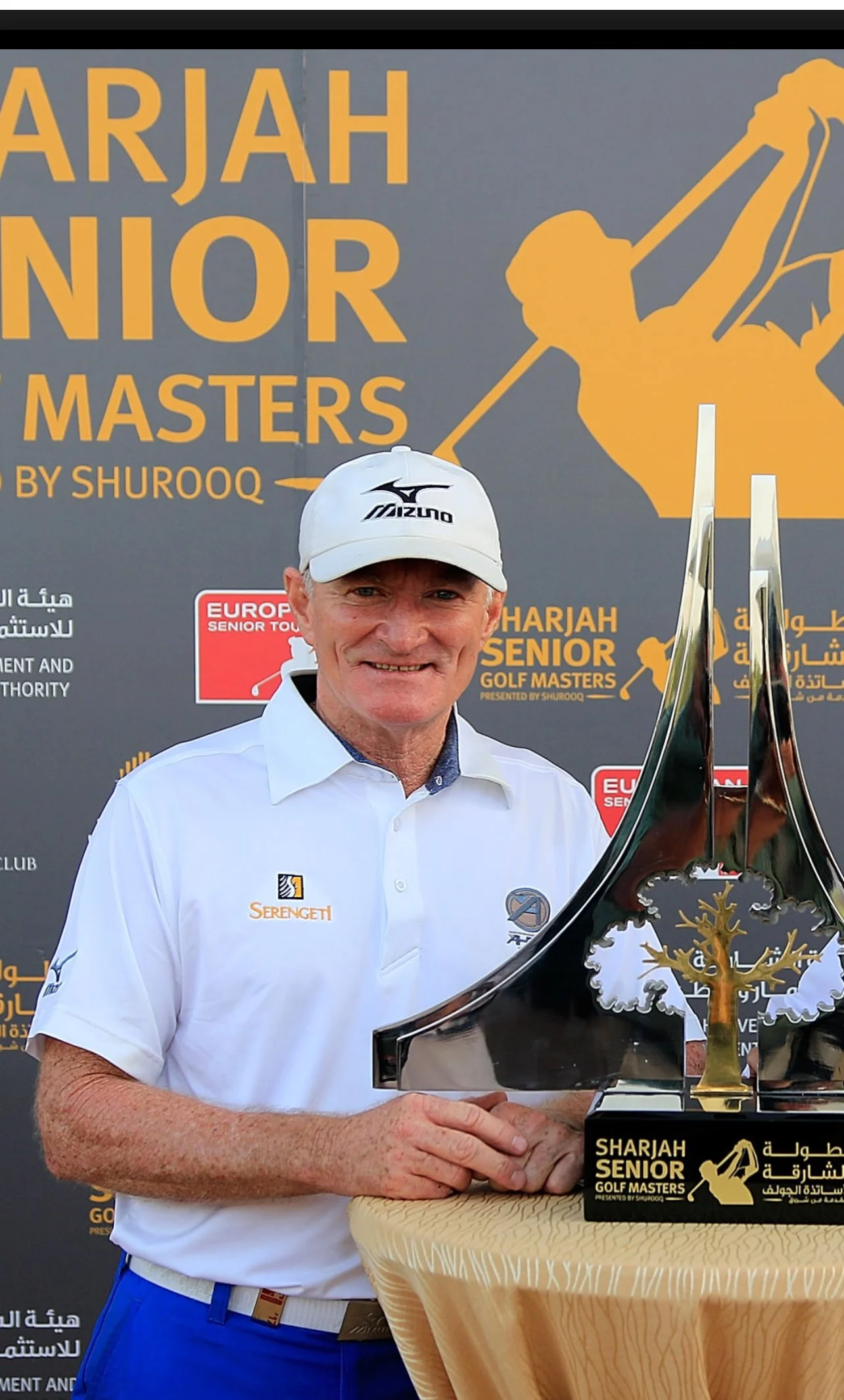 A man in a white polo shirt and cap holding a trophy at the Shariah Senior Golf Masters event.