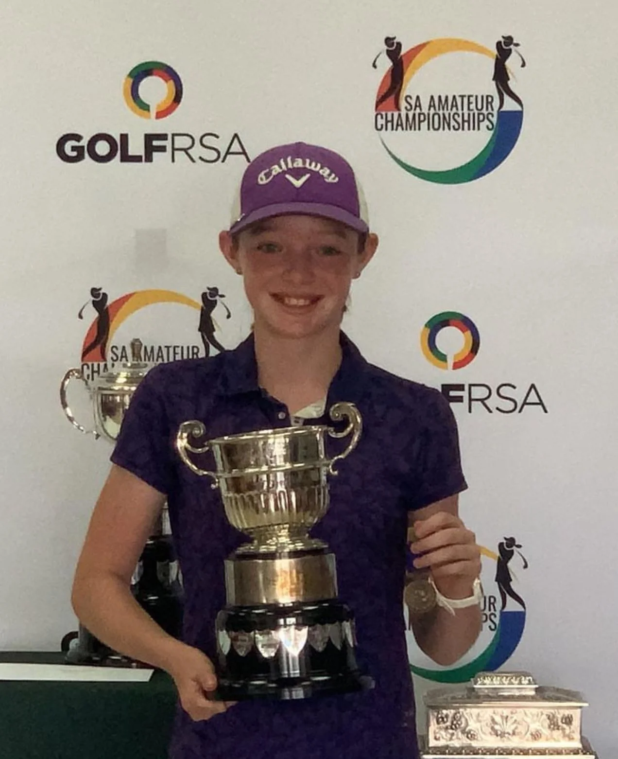 A young girl smiling and holding a large trophy, wearing a purple Callaway cap, at a golf awards ceremony with logos of GOLFRSA and SA Amateur Championships in the background.