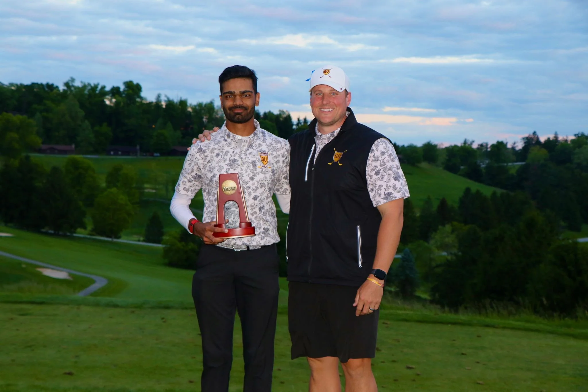 Two men standing on a golf course at sunset. The man on the left is holding a trophy, and the man on the right is wearing a golf hat and vest. Both are smiling.